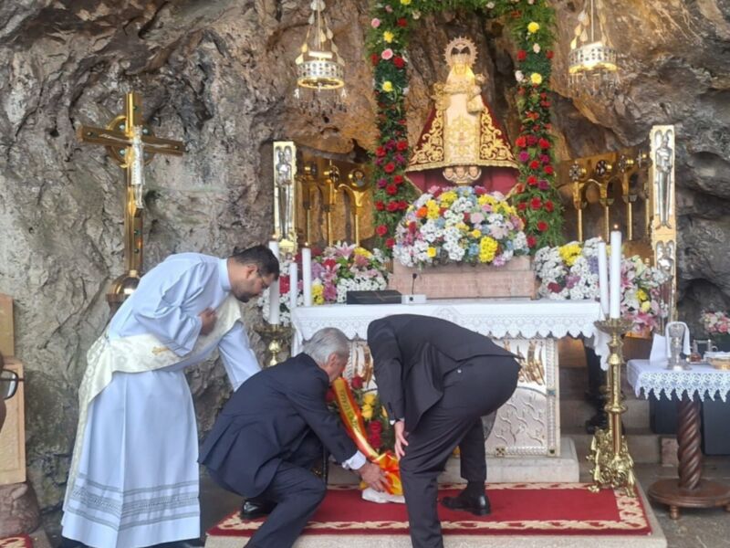 En una cova rocosa, tres personas rezan ante un altar adornado con flores y una imagen religiosa. La escena transmite seriedad y devoción en un ambiente de piedra natural.