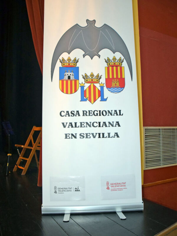 Un cartel blanco con un escudo de armas y el texto "Casa Regional Valenciana en Sevilla" en una sala con asientos.