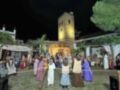 Escena festiva nocturna con una representación de la Natividad en un entorno tradicional, incluyendo figuras vestidas con trajes de época y una estrella en el cielo. La imagen muestra a personas participando activamente en la representación, con una torre de madera al fondo. La escena transmite un ambiente festivo y religioso, probablemente durante la Navidad.