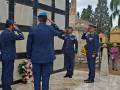 Tres soldados en uniforme militar realizan el saludo de la mano derecha a la estatua de un soldado caído en una tumba conmemorativa. La escena se desarrolla en un entorno de cemento y piedra, con árboles y una estructura arquitectónica en el fondo.