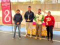 Premiación en un torneo de pádel, con cuatro personas posando en el podio. La imagen muestra trofeos y un banner de la localidad.