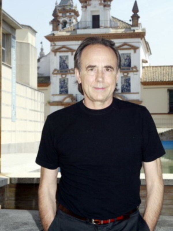 Un hombre con cabello corto y una sonrisa está en el centro de la imagen, con un fondo que incluye una estructura histórica y edificios modernos.