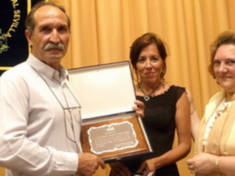 Un hombre sostiene un reconocimiento mientras dos mujeres lo observan. La imagen parece ser de una ceremonia formal, posiblemente un evento académico o empresarial.