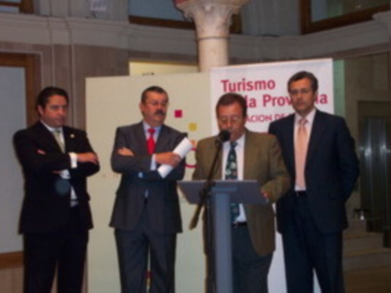 Cuatro hombres en trajes, uno de ellos con corbata roja, se encuentran en un escenario. El hombre del medio sostiene una placa con la palabra "Turismo" y el nombre de la provincia en ella. El fondo muestra un póster con información sobre turismo.
