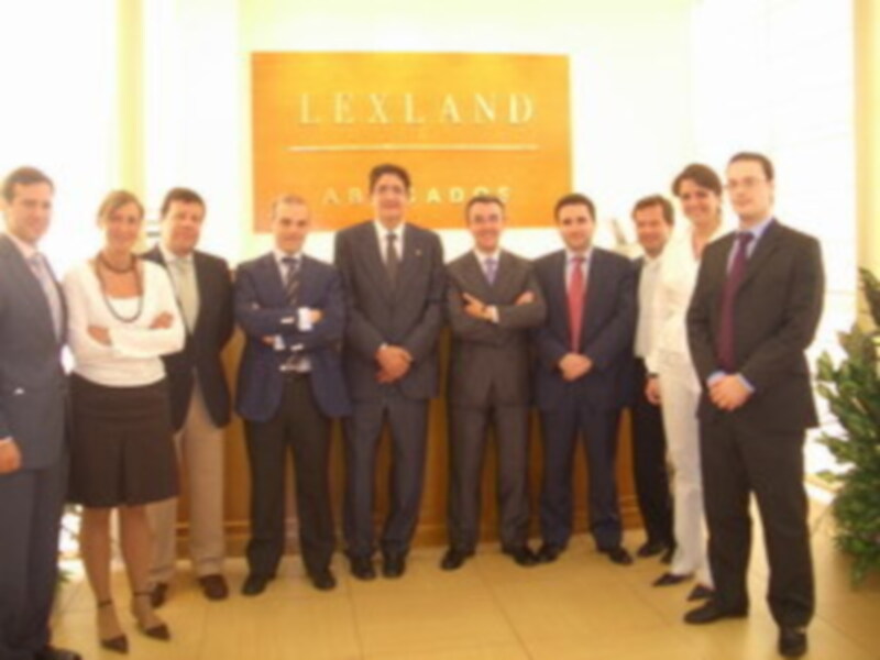 Una oficina con un letrero de Lexland en el fondo. Un grupo de hombres y mujeres están posando, todos con trajes formales. La imagen muestra una reunión o presentación profesional en un entorno de negocios.