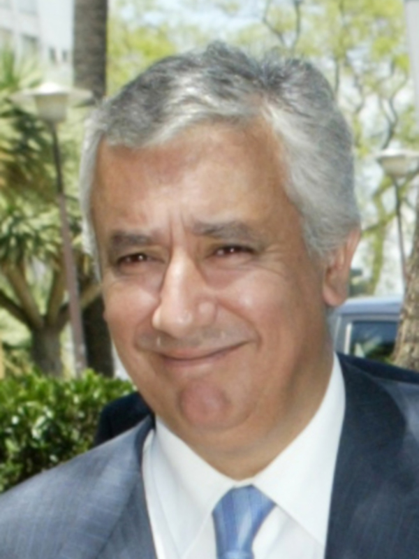 Un hombre con cabello gris, vistiendo una chaqueta azul con una corbata azul claro, posando frente a un árbol y una farola.