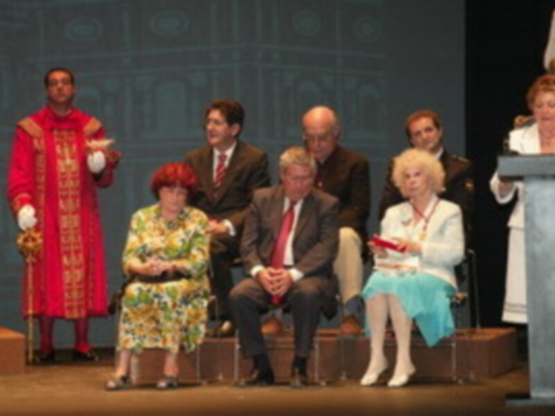 Una escena de un evento formal con personas vestidas formalmente. Un hombre en rojo, una mujer en verde floral, y otros individuos en trajes y vestidos, posan para la cámara en un escenario con un fondo de luces y decoración.