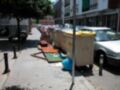 En el paseo de la ciudad, una calle con varios vehículos aparcados y un contenedor de basura en el centro. El área parece desordenada, con restos de papel y otros residuos alrededor del contenedor.