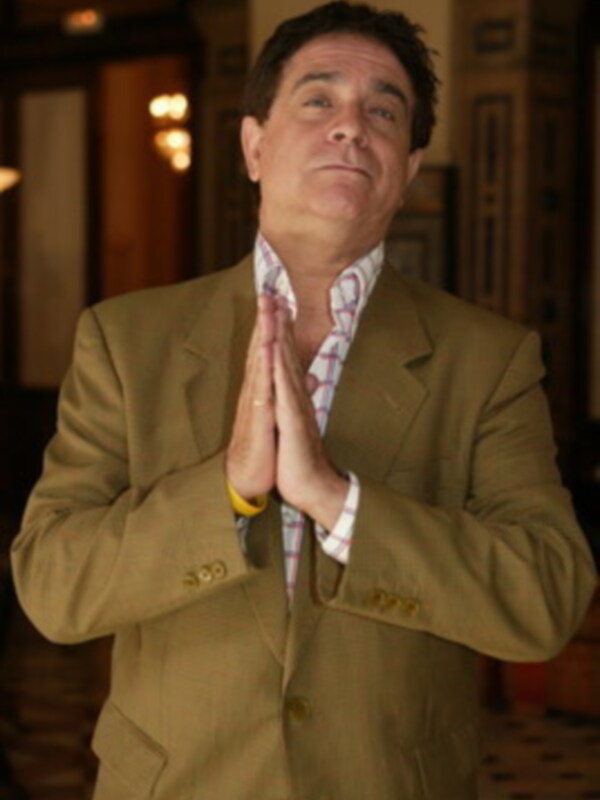 Un hombre con un traje marrón y camisa roja, con las manos en la oración. Fondo de interiores con luces y decoración.