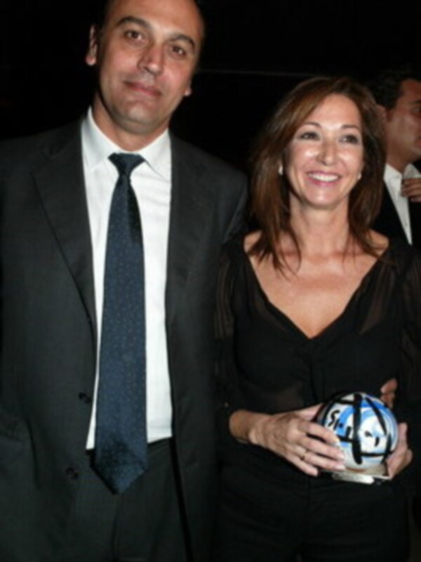 A una pareja en un evento formal. La mujer, vestida con un vestido negro, sostiene un balón de fútbol blanco y azul. El hombre, con una camisa blanca y corbata azul, la mira sonriendo. Ambos parecen estar en una celebración o evento deportivo.