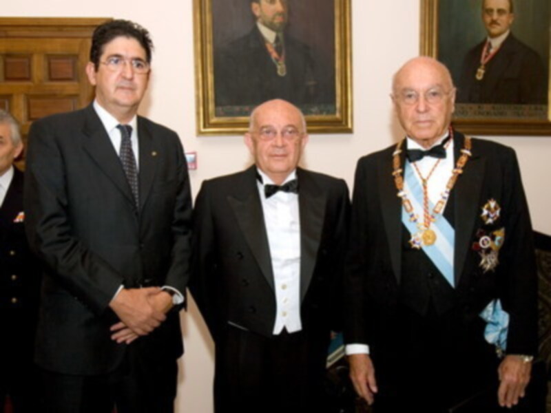Tres hombres vestidos formalmente, posando en una sala con retratos de personas en el fondo.