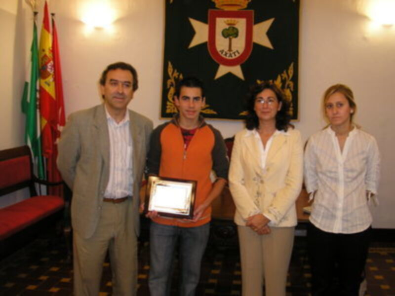 Un joven sostiene un premio mientras se posa junto a dos personas en una sala con banderas y un escudo en el fondo.