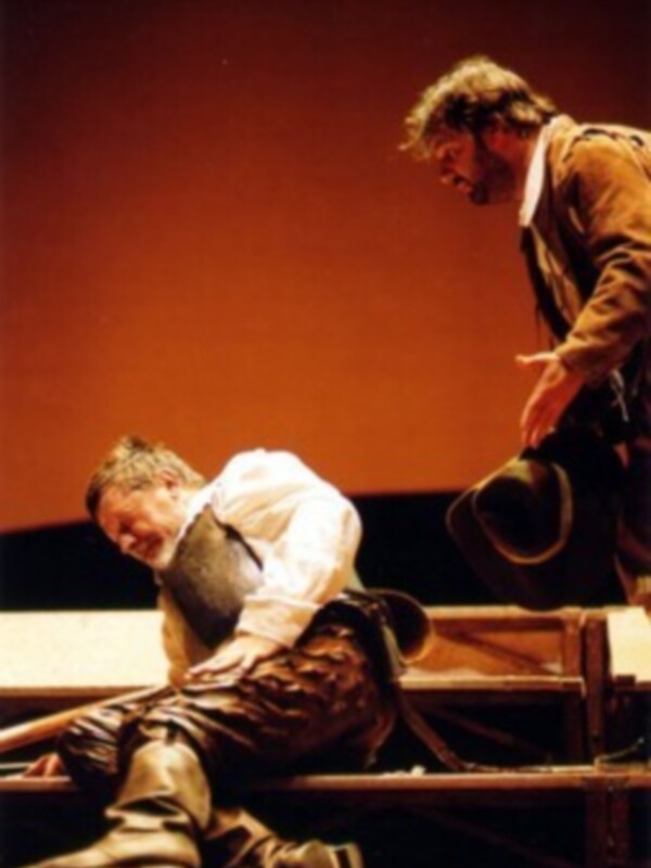 Dos actores en escena, uno sentado y otro de pie, con un fondo teatral rojo.