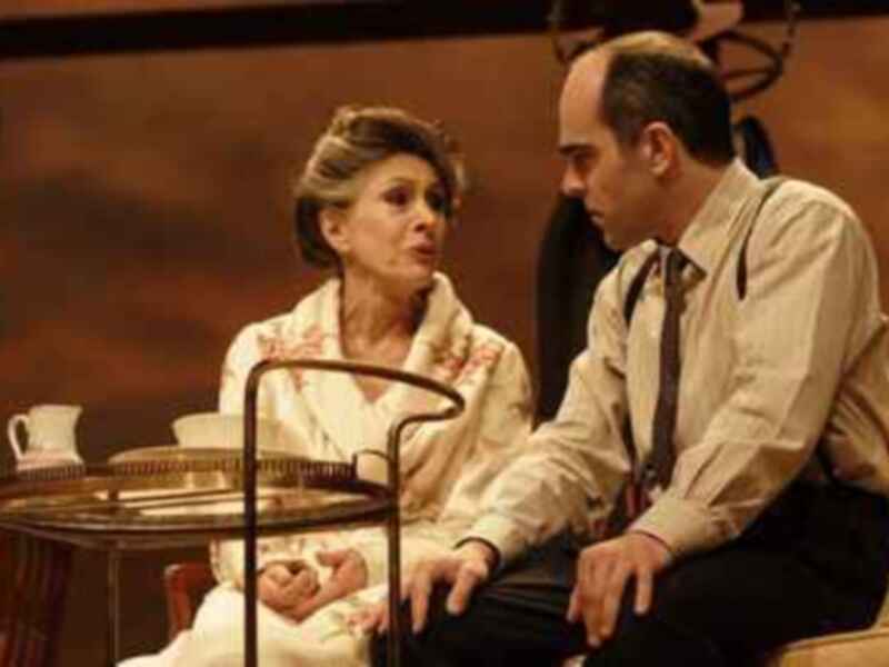 Una escena teatral con una mujer en una camilla y un hombre sentado al lado, ambos mirándose con expresiones serias.