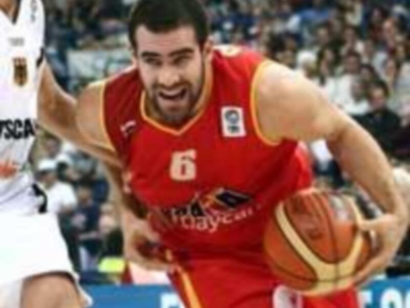 Un jugador de baloncesto en acción durante un partido, con el número 6 y la bandera de España en su camiseta. El fondo muestra a otros jugadores y espectadores en un estadio lleno de público.