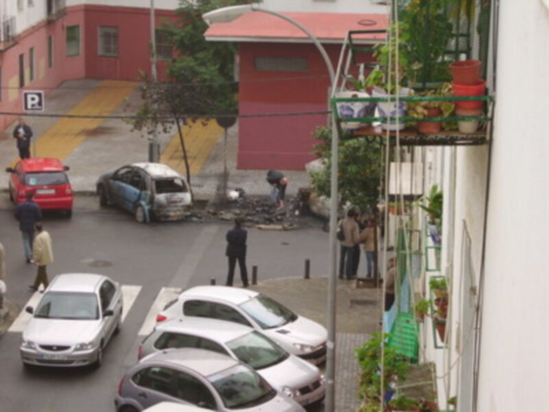 Un coche rojo estacionado en la calle, frente a un edificio rosa con balcones. En el fondo, hay una persona trabajando en la calle y otros vehículos estacionados. La imagen parece ser de un barrio residencial con edificios bajos y una calle tranquila.
