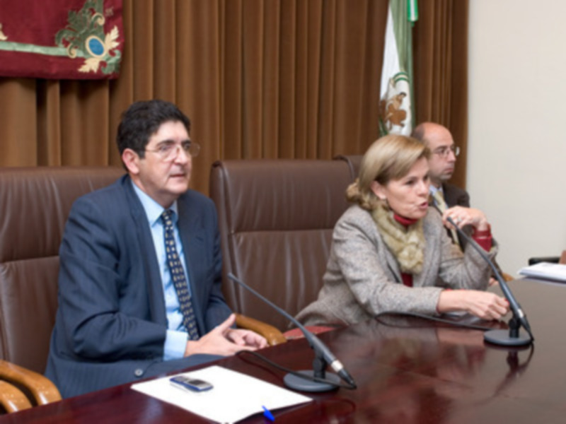 Tres personas sentadas en una mesa de conferencia, uno con un traje y corbata, otra con bufanda y el tercero con una camisa roja. En la mesa hay un teléfono móvil, una pluma y un papel. Detrás de ellos se ven cortinas marrones y banderas.