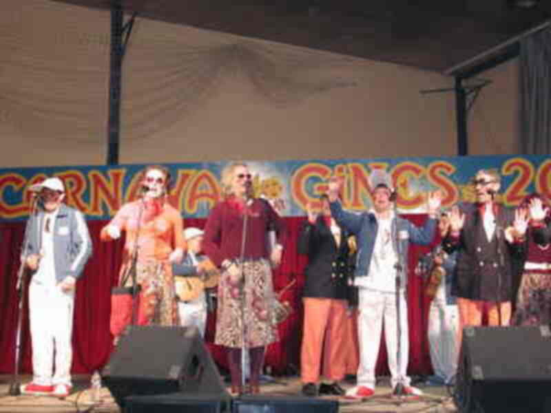 Un grupo de músicos en trajes vintage y sombreros, posiblemente de una banda de música country o rock and roll, se encuentra en un escenario con pancartas que indican "Carnaval de Gines 2013". La escena transmite una celebración festiva y musical.
