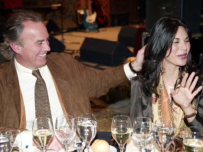 Dos personas sonrientes en un evento formal, uno con una camisa blanca y corbata, la otra con una chaqueta marrón. La imagen muestra detalles de un evento con luces y asistentes en el fondo.