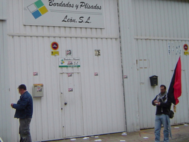 Dos hombres frente a una fábrica con un letrero que dice "Bordados y Plisados León S.L.".