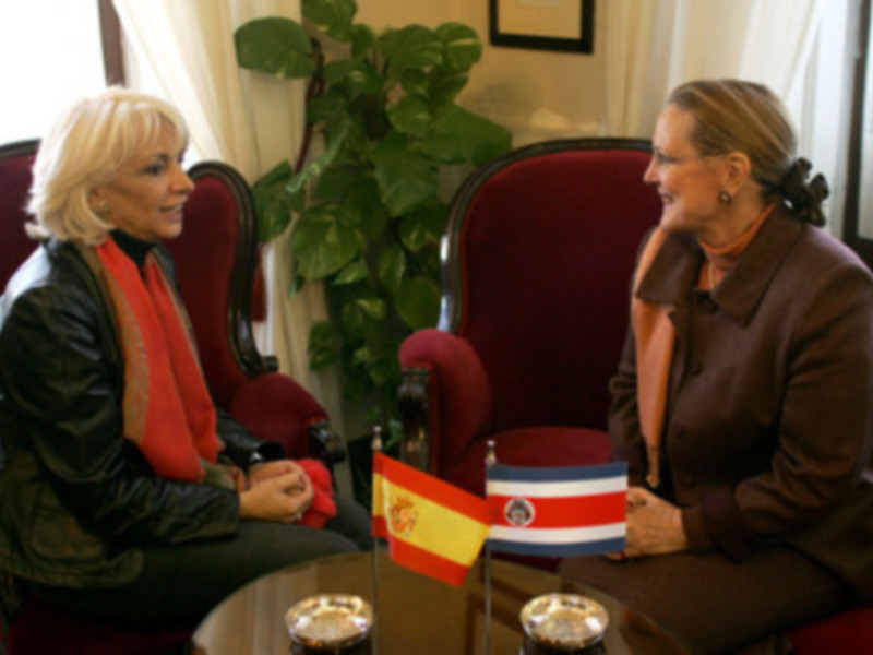 Dos personas charlan en una sala con banderas de España y Costa Rica. La decoración incluye plantas y cortinas blancas.