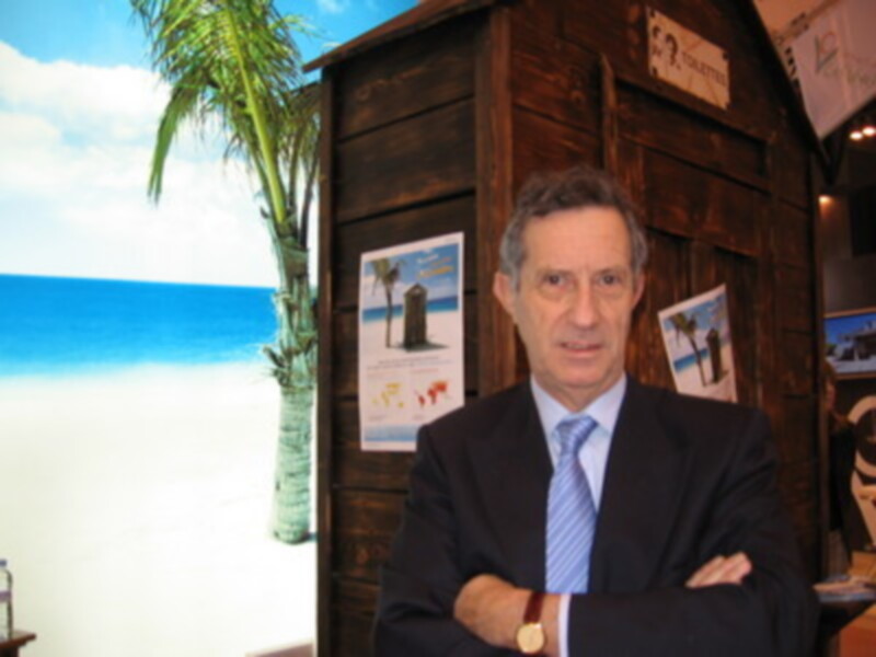 Un hombre en traje y corbata posa frente a una réplica de un bote de madera con una imagen de la playa en el fondo. La escena parece ser parte de un evento o exposición relacionado con la playa y el turismo.