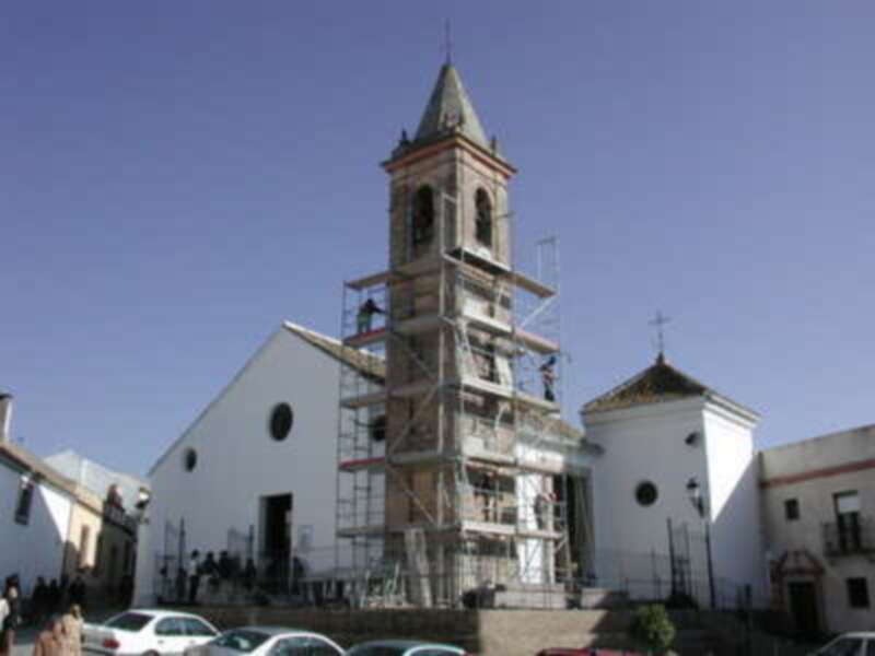 Una iglesia con un campanario en construcción, rodeada por edificios blancos y una calle con vehículos. El cielo es azul claro, indicando buen tiempo.