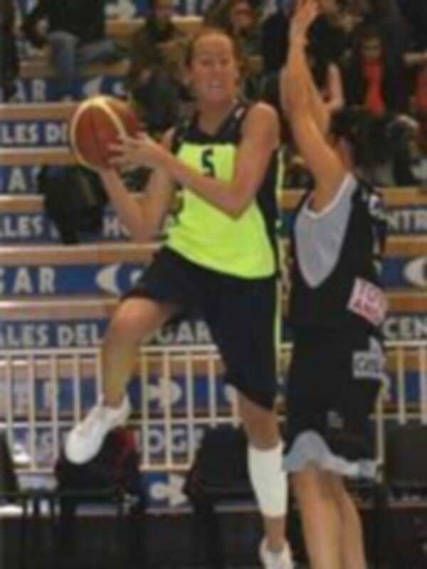 Una jugadora de baloncesto en acción, con el balón en la mano izquierda y la derecha levantada para defender. El fondo muestra un escenario de baloncesto con marcas y publicidad.