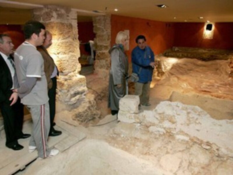 Los visitantes observan una estructura arqueológica en un museo, con paredes de piedra y suelo cubierto de tierra.