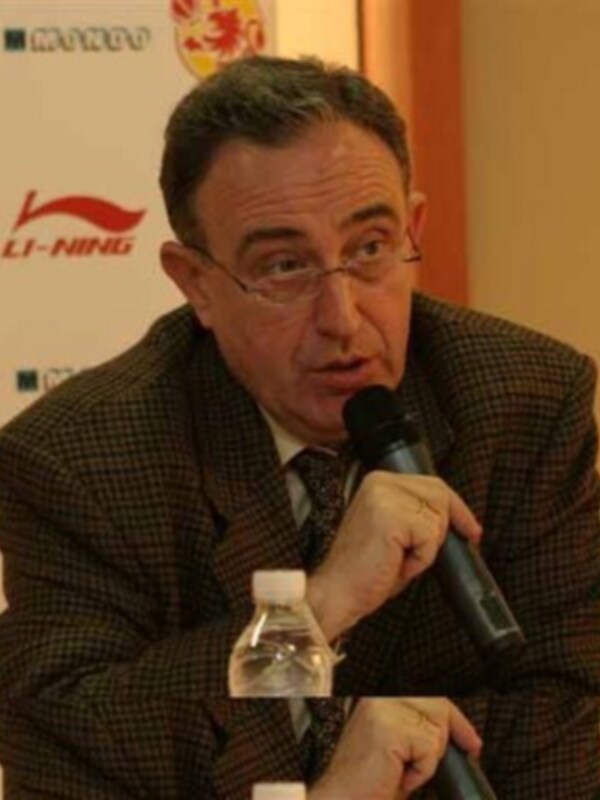 El hombre está hablando en una conferencia de prensa, con un microfono en la mano y una botella de agua a su lado. La pared detrás de él tiene logos de Li-Ning y otras marcas deportivas.
