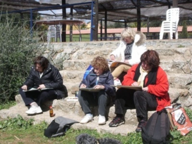 Tres personas sentadas en una banca de piedra, con un bebé entre ellas. La imagen muestra una escena familiar en un parque o jardín, con sombrillas y sillas al fondo.