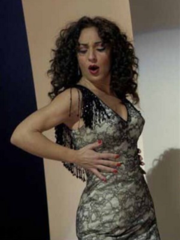 A una mujer con cabello largo y rizado, vestida con un vestido de tirantes de encaje plateado. La postura y expresión sugieren que está en el medio de una actuación o presentación.
