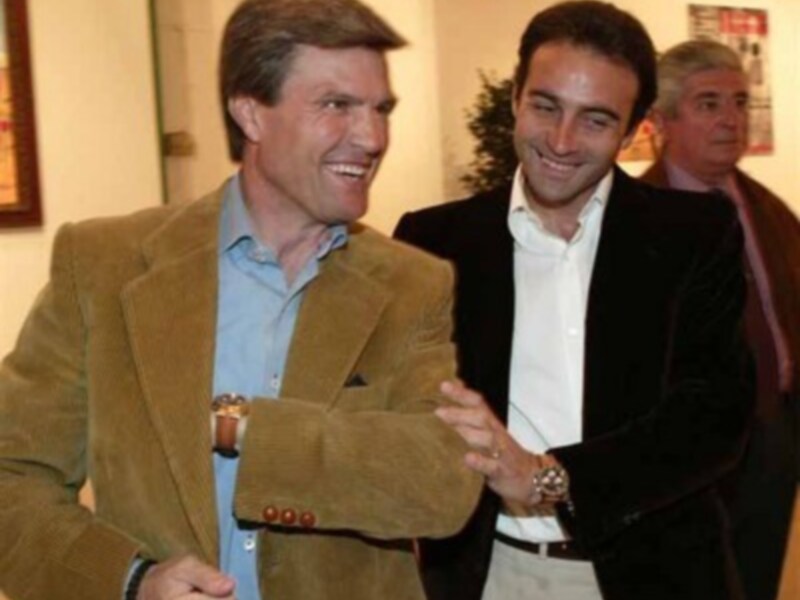 Dos hombres están sonriendo y saludándose en una sala con decoración formal.