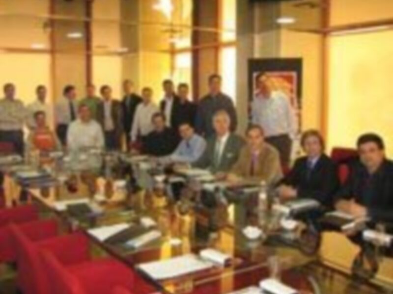 Una reunión formal en una sala con mesas redondas, sillas rojas y un televisor. Personas sentadas en grupos conversando, con una mesa central que parece tener documentos y un teléfono. Ambiente profesional y organizado.