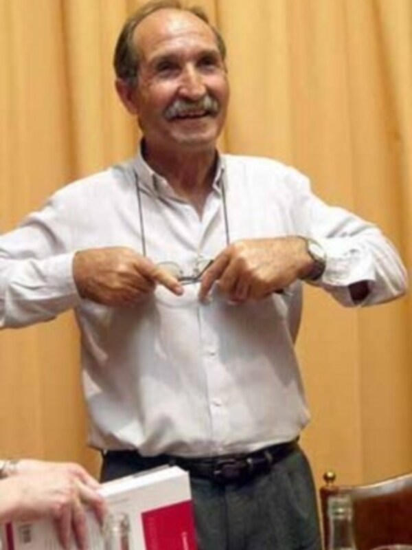 Un hombre con barba y bigote sonríe mientras sostiene un libro abierto en una mano. La imagen sugiere que está presentando o discutiendo el contenido del libro con otra persona a la izquierda.