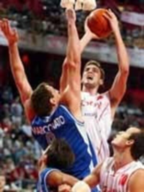 Un jugador de baloncesto en un partido, con una camiseta blanca y un jersey azul sobre la cabeza. El fondo muestra a otros jugadores y una grada llena de espectadores.
