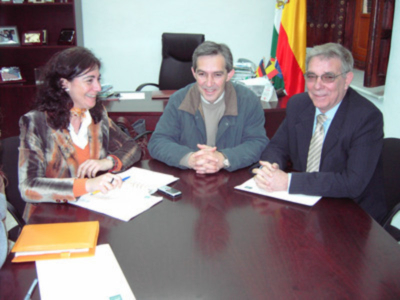 Tres personas sentadas en una mesa, con un documento en el centro, en un entorno formal con una bandera en el fondo.