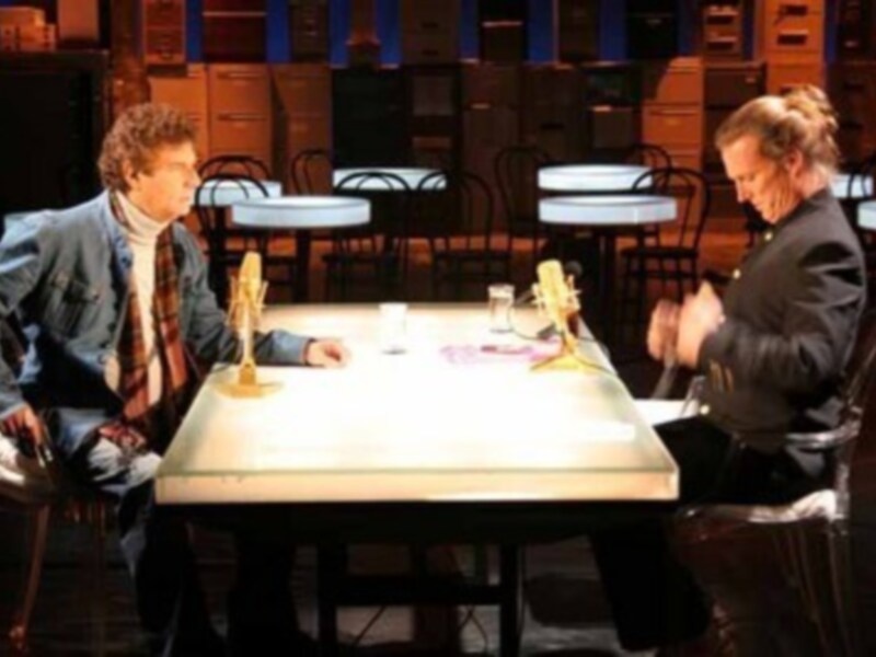 Duo de actores en una escena de teatro, sentados en una mesa con microfonos, en un escenario iluminado.
