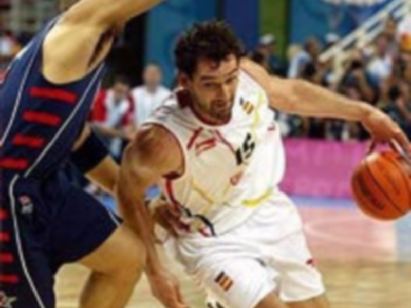 Un jugador de baloncesto en acción, con una camiseta blanca y roja, un balón en la mano izquierda, y otro jugador de equipo contrario a su derecha. El fondo muestra un estadio lleno de espectadores y una publicidad en el muro.