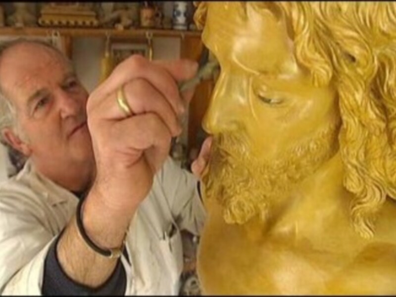 Un artista trabajando en una escultura dorada de un personaje con barba y cabello largo, usando un pincel para aplicar detalles finos.