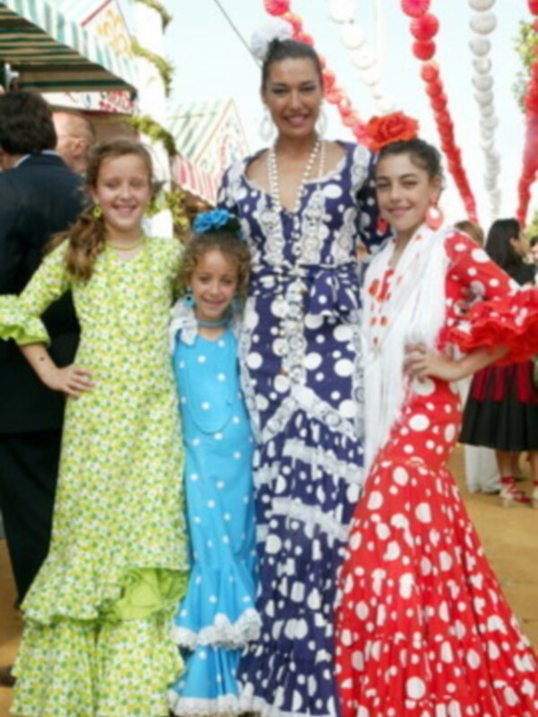 Tres niñas vestidas con trajes tradicionales españoles posan para una foto en un ambiente festivo. La imagen muestra detalles de la ropa y el entorno decorado con globos y estandartes.