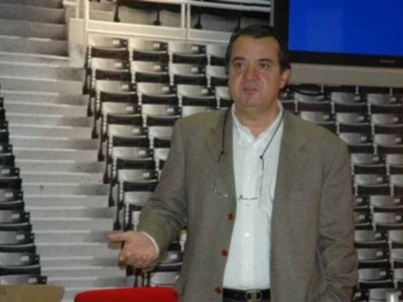 Un hombre en traje beige y camisa blanca se encuentra frente a una pantalla de televisión, con un fondo de asientos en gradas.