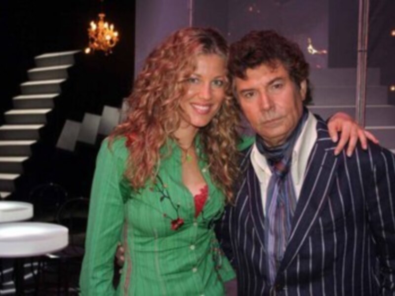 A una mujer con cabello rojo largo y una blusa verde brillante, posando junto a un hombre con una camisa azul y traje de rayas. Ambos están en un escenario con escaleras blancas detrás, iluminado por luces doradas.