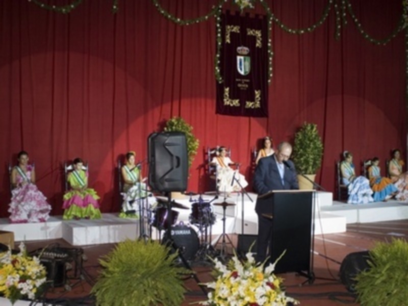 Una escena formal con una mujer en un vestido rosa sentada y otros vestidos de colores en el fondo. Un hombre está al frente con un micrófono y un altavoz. Detalles decorativos como flores y una bandera en el fondo.