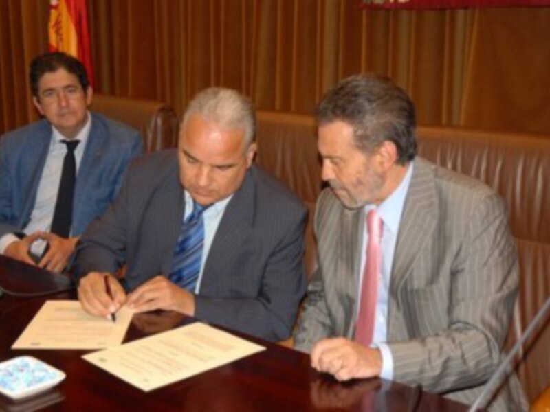 Dos hombres en trajes firman documentos en una sala de conferencias. El ambiente es formal, con sillas y una bandera visible al fondo.