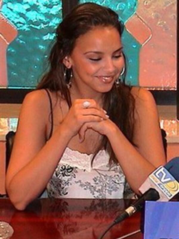 A una mujer con cabello largo y ondulado, llevando un vestido blanco con flores. Está sentada en una mesa de conferencia, con un micrófono y un logo visible en el fondo. La escena parece ser una entrevista o presentación formal.