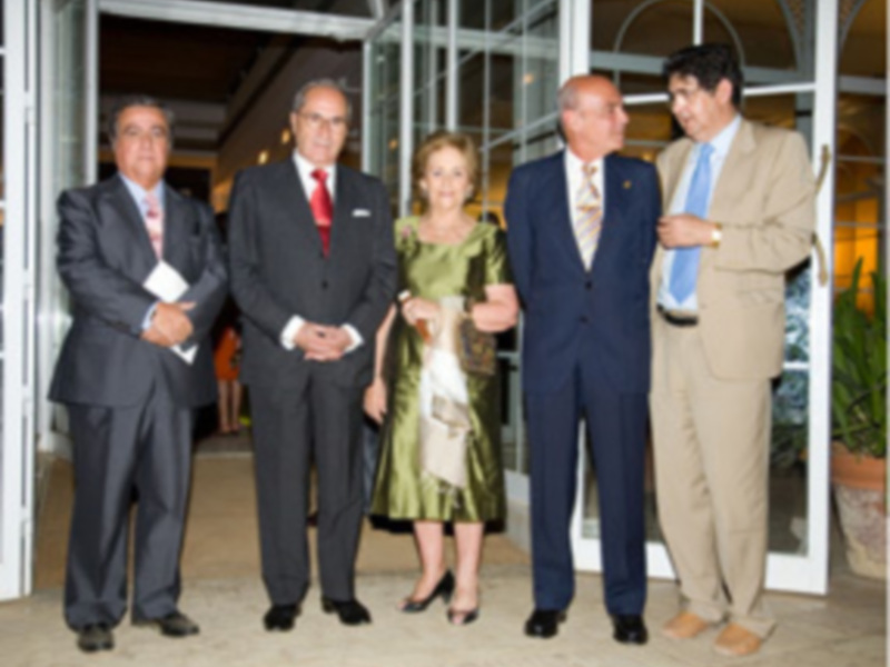 Un grupo de personas vestidas formalmente, posando frente a una estructura metálica. La imagen parece ser de un evento formal o ceremonial, posiblemente en una celebración o conferencia.