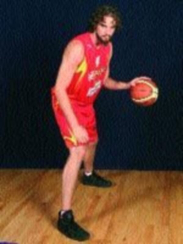 Un jugador de baloncesto en acción, con una camiseta roja y amarilla, pantalones cortos rojos y zapatillas negras. El suelo es de madera, probablemente en un campo de baloncesto.