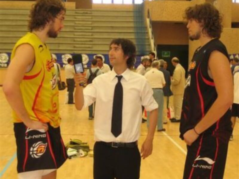 Un hombre con una camiseta amarilla y negro, otro con una camiseta blanca y negro y el tercero con una camiseta negra y roja. Todos están en un gimnasio, posiblemente antes de un partido o entrenamiento.