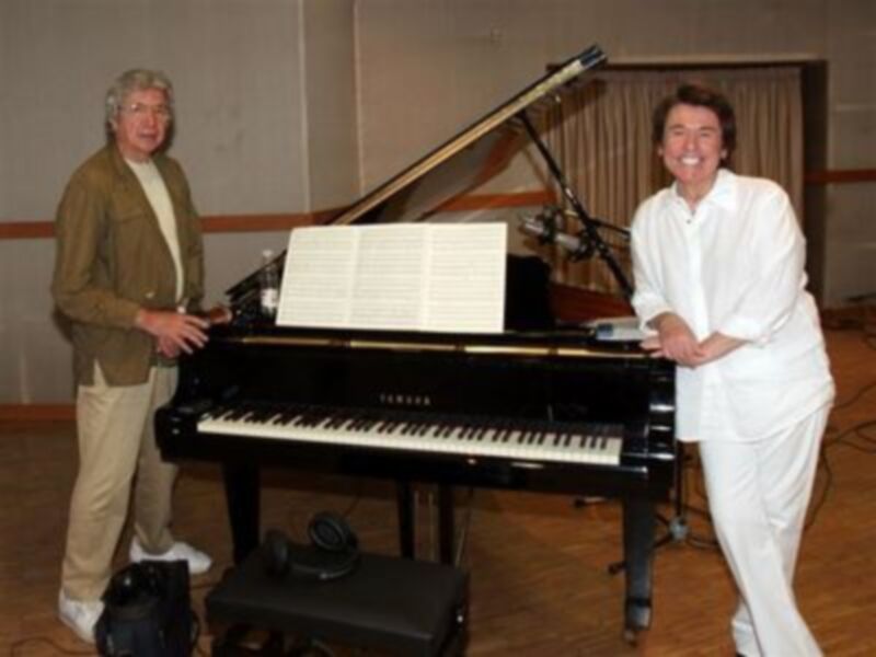 Dos personas posan junto a un piano en un entorno acústico, posiblemente en un estudio de grabación o sala de ensayo.